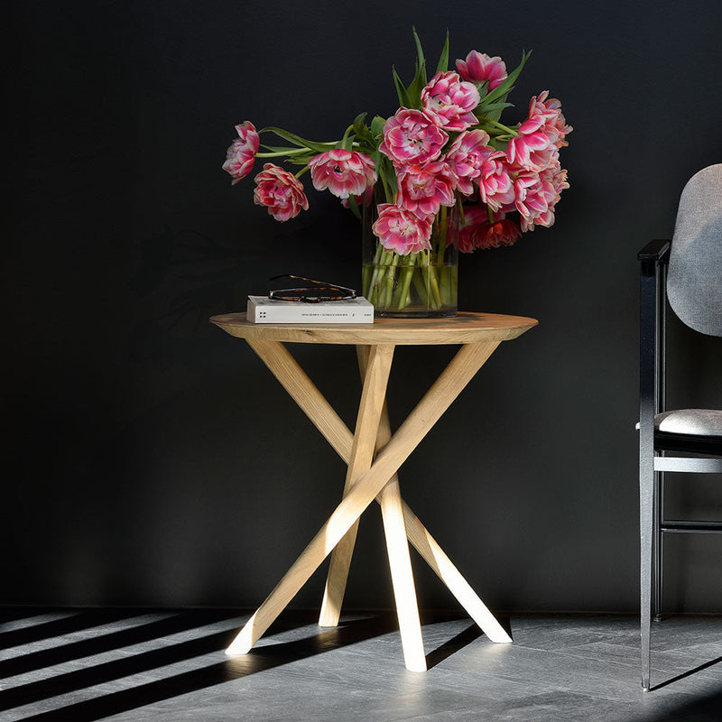 elements solid oak side table with round top and crossed  legs, shown with vase of flowers on top.