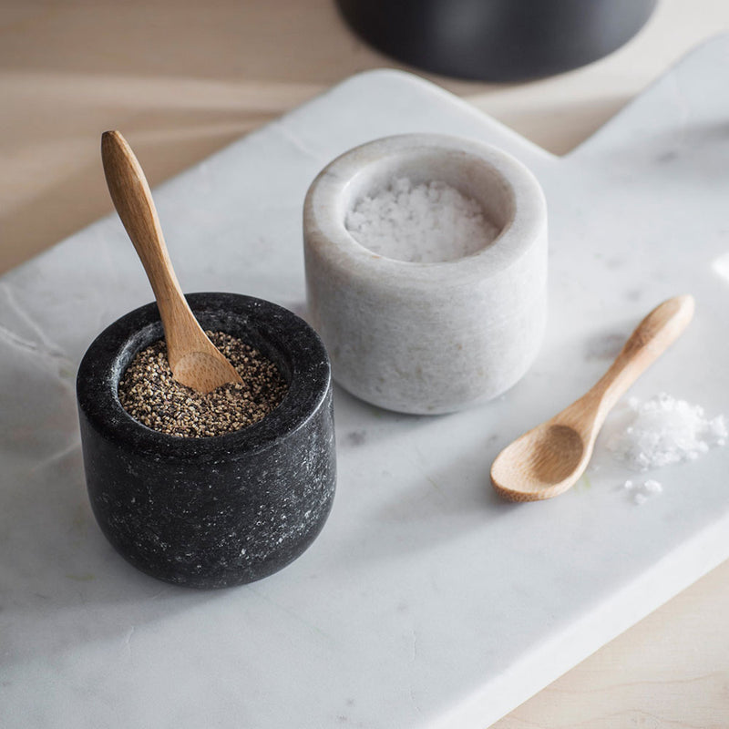 pair of pinch pots, one light marble, one dark granite with wooden spoons, shown with salt and pepper in.
