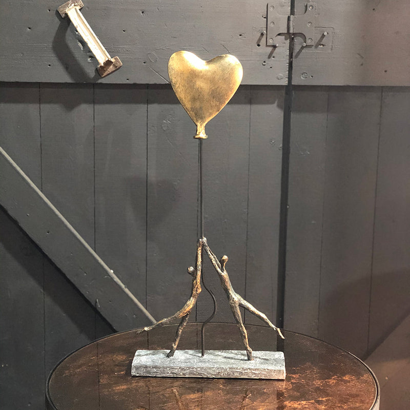 a bronze colour sculpture of two people hoding on to the rope of a large gold heart shaped balloon.